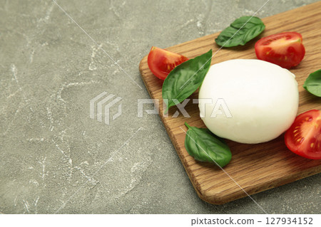 Mozzarella cheese with green basil leaves and tomato on cutting board on grey background Mozzarella cheese with green basil leaves and tomato on cutting board on grey background 127934152