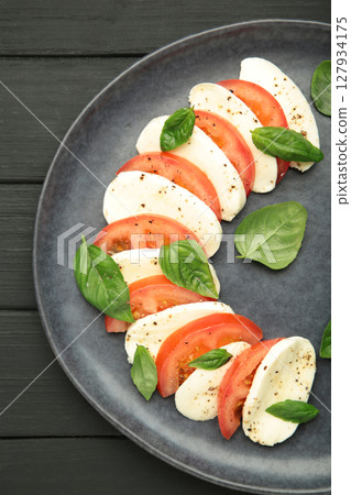 Italian Caprese salad with cherry tomatoes, mozzarella, basil on dark plate on black wooden background. Vertical photo Italian Caprese salad with cherry tomatoes, mozzarella, basil on dark plate on black wooden background. Vertical photo 127934175