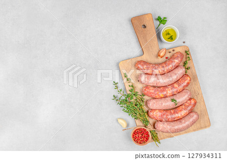 A rustic arrangement of raw sausages with herbs, oil, and spices on a wooden cutting board over a clean light background, perfect for culinary and cooking concepts 127934311