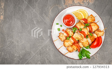Grilled chicken skewers served with fresh vegetables, herbs, and tomato sauce on a rustic plate over a dark stone background 127934313
