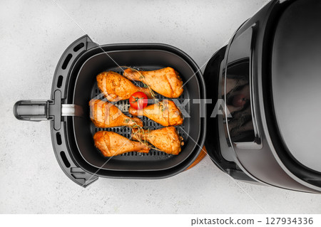 Top view of seasoned chicken drumsticks and a tomato cooking inside a modern black air fryer on a light kitchen counter Top view of seasoned chicken drumsticks and a tomato cooking inside a modern black air fryer on a light kitchen counter 127934336