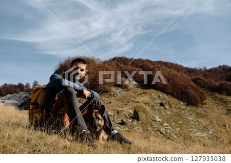 A young man resting with his German Shepherd dog in the golden grass of Prokletije National Park, Montenegro, surrounded by autumn forest and rocky mountain slopes. Hiking with pet concept 127935038