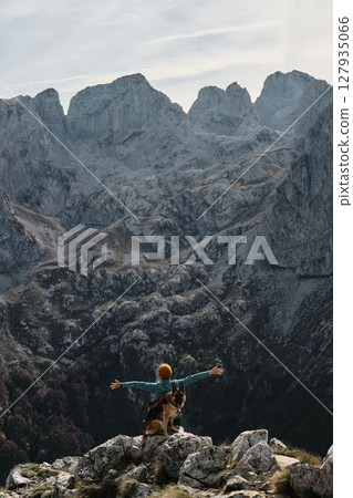 A female traveler raises her arms in triumph while standing with her German Shepherd atop a rocky ridge in Prokletije National Park, Montenegro. Rear view. Travel with pet concept A female traveler raises her arms in triumph while standing with her German Shepherd atop a rocky ridge in Prokletije National Park, Montenegro. Rear view. Travel with pet concept 127935066