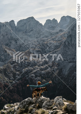 A female traveler raises her arms in triumph while standing with her German Shepherd atop a rocky ridge in Prokletije National Park, Montenegro. Rear view. Travel with pet concept A female traveler raises her arms in triumph while standing with her German Shepherd atop a rocky ridge in Prokletije National Park, Montenegro. Rear view. Travel with pet concept 127935067