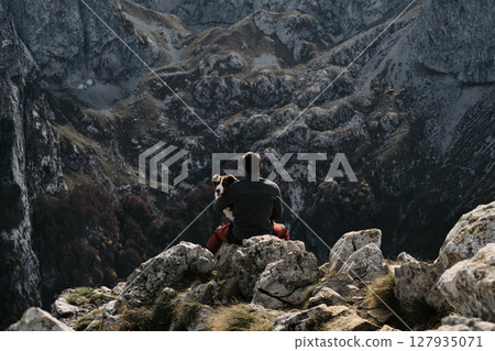 Sitting on rocky cliffs, a man embraces his Australian Shepherd while overlooking Montenegro majestic Prokletije range in autumn. Hiking with pet concept Sitting on rocky cliffs, a man embraces his Australian Shepherd while overlooking Montenegro majestic Prokletije range in autumn. Hiking with pet concept 127935071