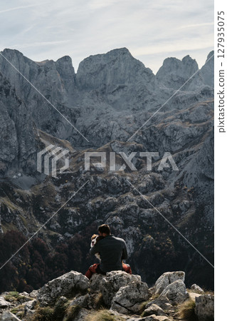 Sitting on rocky cliffs, a man embraces his Australian Shepherd while overlooking Montenegro majestic Prokletije range in autumn. Hiking with pet concept Sitting on rocky cliffs, a man embraces his Australian Shepherd while overlooking Montenegro majestic Prokletije range in autumn. Hiking with pet concept 127935075