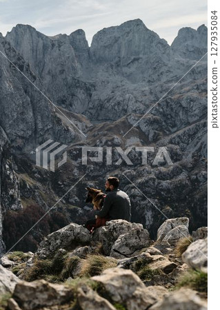 A man sits on rocky terrain, hugging his German Shepherd dog with towering mountains of Prokletije in Montenegro behind them. Rear view. Hiking with pet concept A man sits on rocky terrain, hugging his German Shepherd dog with towering mountains of Prokletije in Montenegro behind them. Rear view. Hiking with pet concept 127935084