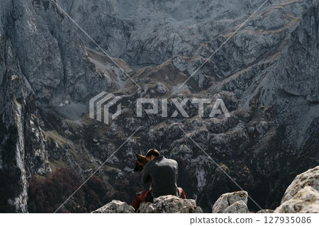 A man sits on rocky terrain, hugging his German Shepherd dog with towering mountains of Prokletije in Montenegro behind them. Rear view. Hiking with pet concept 127935086