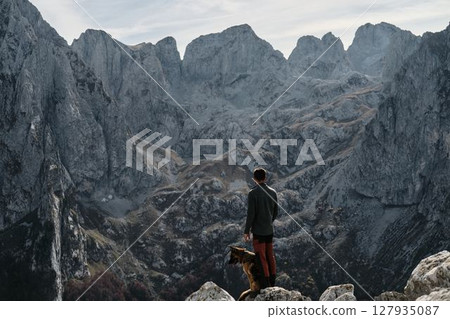 A man and his German Shepherd stand on a mountain edge in Montenegro, Prokletije National Park, taking in the vast alpine view during a crisp fall day. Rear view. Hiking with pet concept A man and his German Shepherd stand on a mountain edge in Montenegro, Prokletije National Park, taking in the vast alpine view during a crisp fall day. Rear view. Hiking with pet concept 127935087