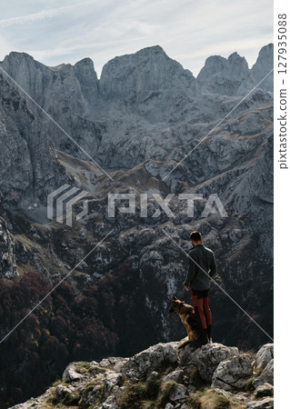 A man and his German Shepherd stand on a mountain edge in Montenegro, Prokletije National Park, taking in the vast alpine view during a crisp fall day. Rear view. Hiking with pet concept A man and his German Shepherd stand on a mountain edge in Montenegro, Prokletije National Park, taking in the vast alpine view during a crisp fall day. Rear view. Hiking with pet concept 127935088