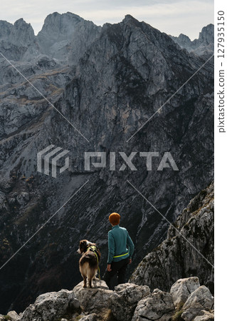 A woman and her Australian shepherd dog enjoy the majestic mountain view from a rocky ridge in Prokletije National Park, Montenegro, during fall. Rear view. Hiking with pet concept A woman and her Australian shepherd dog enjoy the majestic mountain view from a rocky ridge in Prokletije National Park, Montenegro, during fall. Rear view. Hiking with pet concept 127935150