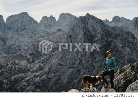 A female hiker stands proudly on a rocky cliff with her Australian Shepherd dog, framed by the dramatic peaks of Prokletije, Montenegro in autumn season. Hiking with pet concept 127935153