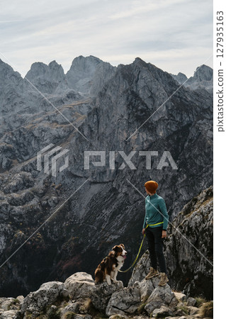 A female hiker stands proudly on a rocky cliff with her Australian Shepherd dog, framed by the dramatic peaks of Prokletije, Montenegro in autumn season. Hiking with pet concept 127935163