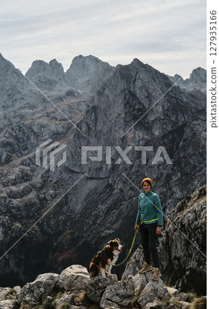 A female hiker stands proudly on a rocky cliff with her Australian Shepherd dog, framed by the dramatic peaks of Prokletije, Montenegro in autumn season. Hiking with pet concept A female hiker stands proudly on a rocky cliff with her Australian Shepherd dog, framed by the dramatic peaks of Prokletije, Montenegro in autumn season. Hiking with pet concept 127935166