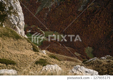 Tiny remote houses sit far below in a green meadow surrounded by dense autumn forest and steep cliffs in Prokletije. Aerial view Tiny remote houses sit far below in a green meadow surrounded by dense autumn forest and steep cliffs in Prokletije. Aerial view 127935175