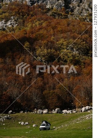 White camper van parked on a green meadow at the edge of colorful autumn forest below the mountains in Prokletije, Montenegro. Vanlife outdoor travel concept 127935200