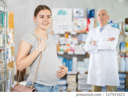 Pregnant woman holding prescription in her hands in interior of modern pharmacy. In background, male pharmacist offers to buy medicine Pregnant woman holding prescription in her hands in interior of modern pharmacy. In background, male pharmacist offers to buy medicine 127935821