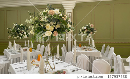 Elegant banquet hall with white tablecloths, floral centerpieces, and napkin arrangements prepared for a formal celebration, wedding, or upscale event reception setup Elegant banquet hall with white tablecloths, floral centerpieces, and napkin arrangements prepared for a formal celebration, wedding, or upscale event reception setup 127937588