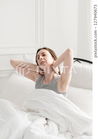 Young and beautiful woman sits on a bed with white bed linen and stretches after sleep. Young and beautiful woman sits on a bed with white bed linen and stretches after sleep. 127940673