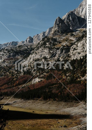 Sharp limestone peaks dominate the skyline above layers of fall-colored trees in Prokletije National Park, Montenegro in autumn season. 127940888