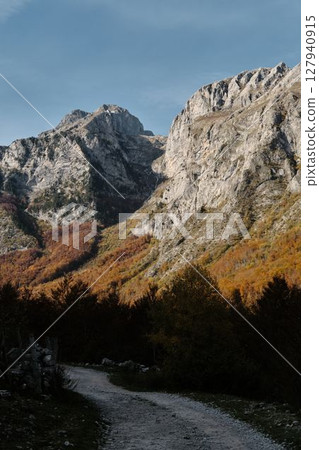 Narrow trail winding through vibrant autumn forest beneath steep cliffs in Prokletije National Park, Montenegro, a peaceful hiking scene 127940915