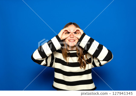 Beautiful Hispanic teenager with braces wearing a striped sweater making a thumbs-up gesture, as if holding binoculars, looking through her fingers. Fun concept Beautiful Hispanic teenager with braces wearing a striped sweater making a thumbs-up gesture, as if holding binoculars, looking through her fingers. Fun concept 127941345