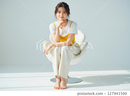 Female model in a white T-shirt and yellow camisole sitting on a white chair against a white background Female model in a white T-shirt and yellow camisole sitting on a white chair against a white background 127941500