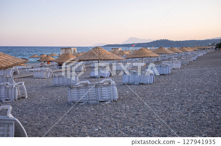 Beach scene with empty sun loungers and straw umbrellas under a pastel sky at sunset Beach scene with empty sun loungers and straw umbrellas under a pastel sky at sunset 127941935