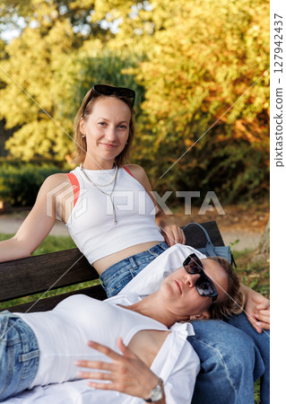 Pair smiling happy young adult women enjoy sunny day together sit relaxing on bench in city park outdoors. Female persons wearing casual denim white tops portrait. Best friends friendship outside 127942437