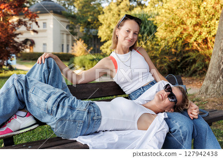 Pair smiling happy young adult women enjoy sunny day together sit relaxing on bench in city park outdoors. Female persons wearing casual denim white tops portrait. Best friends friendship outside 127942438