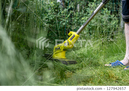Man trimming overgrown grass with electric string trimmer in summer garden 127942657
