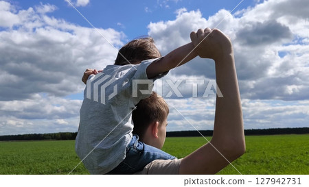 Kids is on brother's shoulders with his arms outstretched. rear view. Boy sitting on brother's shoulders. Happy family. Family vacation, travel in summer. concept of a happy childhood 127942731