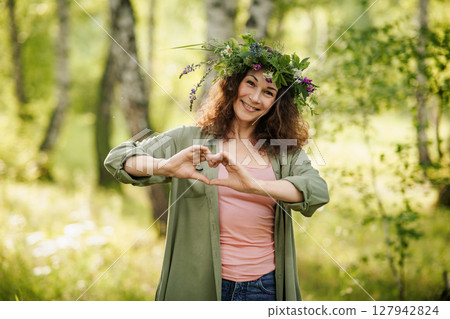 Cheerful woman in floral wreath forming heart gesture with hands in green forest, summer day. Symbol of care, positivity, mindfulness and joy in nature 127942824