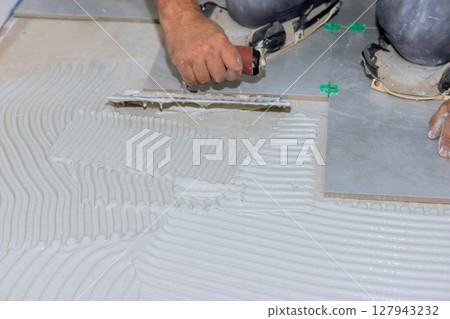 Worker skillfully spreads tile adhesive on floor surface for home renovation project. 127943232