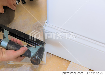 Worker applies finishing touches to home by installing new baseboard trim with nail gun in living room. Worker applies finishing touches to home by installing new baseboard trim with nail gun in living room. 127943238