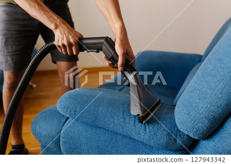 Close-up cropped shot of unrecognizable cleaning service worker using washing vacuum cleaner to clean blue sofa in modern living room. Concept of hygiene and cleanliness in domestic environment. Close-up cropped shot of unrecognizable cleaning service worker using washing vacuum cleaner to clean blue sofa in modern living room. Concept of hygiene and cleanliness in domestic environment. 127943342