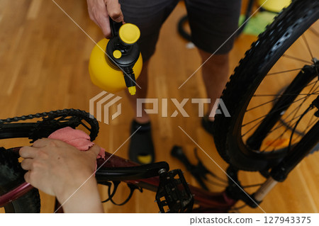Two mechanics male cleaning mountain bike using water sprayer and cloth, removing dirt and grime from frame and chain at home, performing detailed maintenance after trail riding, close-up. Two mechanics male cleaning mountain bike using water sprayer and cloth, removing dirt and grime from frame and chain at home, performing detailed maintenance after trail riding, close-up. 127943375