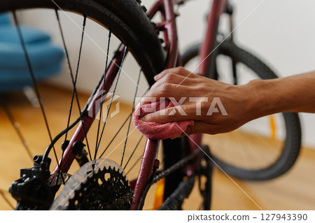 Close-up hands of cyclist male cleaning mountain bike frame with microfiber cloth, performing detailed maintenance after trail riding, preserving cycling gear condition and functionality at home. 127943390