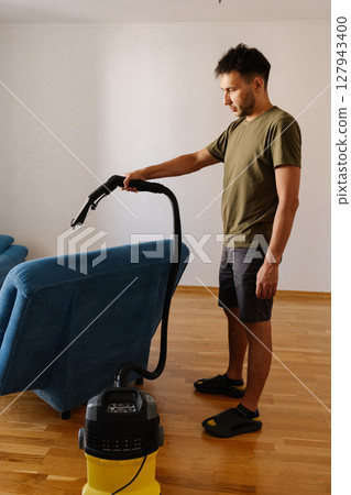 Side view of handsome bearded young man in casual clothes removing dirt and stains using washing vacuum cleaner to clean blue sofa in living room, demonstrating modern approach to household chores. Side view of handsome bearded young man in casual clothes removing dirt and stains using washing vacuum cleaner to clean blue sofa in living room, demonstrating modern approach to household chores. 127943400