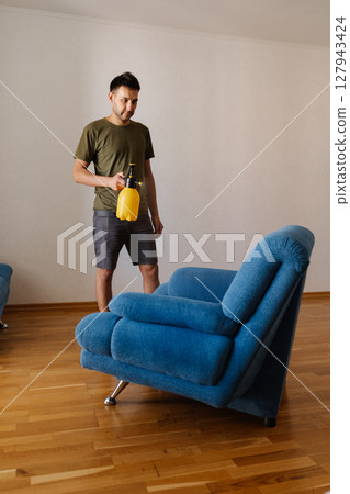 Full length vertical portrait of young man spraying water using yellow plastic sprayer on blue sofa in stylish living room, engaging home cleaning and maintenance. Concept of household cleanliness. Full length vertical portrait of young man spraying water using yellow plastic sprayer on blue sofa in stylish living room, engaging home cleaning and maintenance. Concept of household cleanliness. 127943424