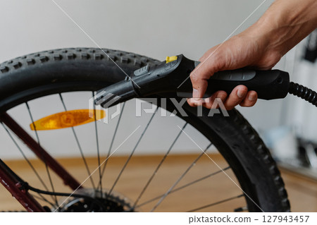 Cyclist male using steam cleaner to remove dirt and dust from reflector of bicycle wheel at home, ensuring cleanliness, close-up. Concept of professional service and maintenance of bike transport. 127943457