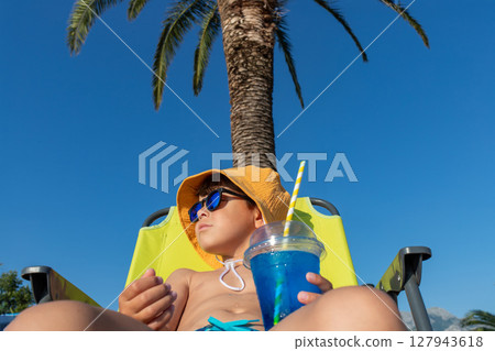 Boy in beachwear relaxing with a drink in the shade of a palm tree 127943618