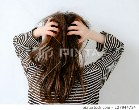 一位身穿條紋印花衣服的女士,手放在長長的棕色頭髮上,表情憂慮而煩惱,背景是一堵牆 一位身穿條紋印花衣服的女士,手放在長長的棕色頭髮上,表情憂慮而煩惱,背景是一堵牆 127944754