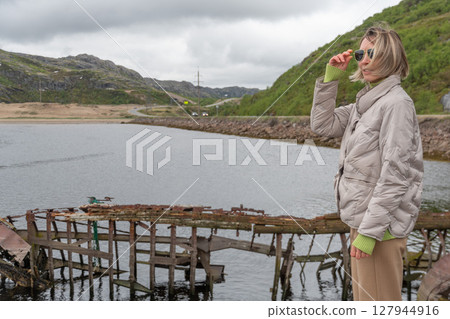 Woman in beige jacket and sunglasses standing by a calm river with an old wooden shipwreck and winding coastal road in the background 127944916