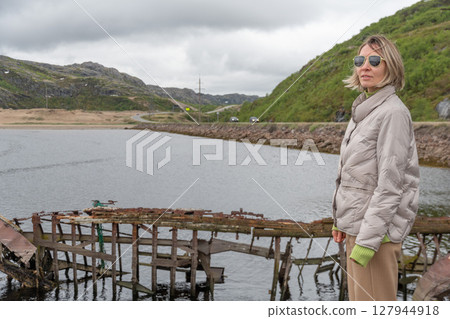 Woman in beige jacket and sunglasses standing by a calm river with an old wooden shipwreck and winding coastal road in the background 127944918