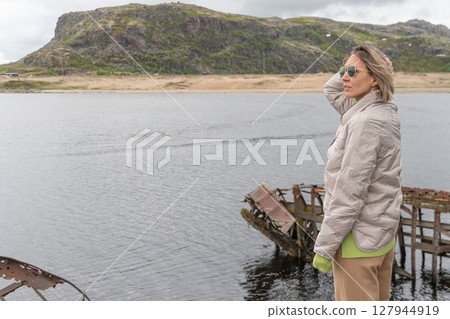 Woman in beige jacket and sunglasses standing by a calm river with an old wooden shipwreck and winding coastal road in the background 127944919