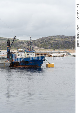 Old blue fishing vessel anchored near the shore with rugged hills in the background and cloudy sky above Old blue fishing vessel anchored near the shore with rugged hills in the background and cloudy sky above 127944931