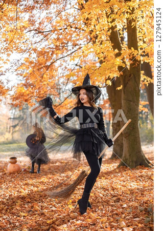 Brunette woman in witch costume standing in autumn forest on Halloween day. Woman wearing black clothes and cone hat. Woman sitting on a broom. 127945124
