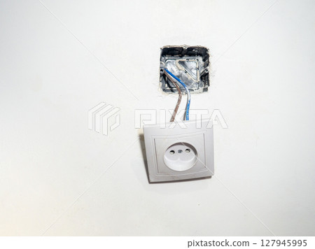 Broken electrical socket on the wall. Disassembled device. White wall with a socket pulled out of the wall. Life threatening. Broken electrical equipment concept. Appliances. 127945995