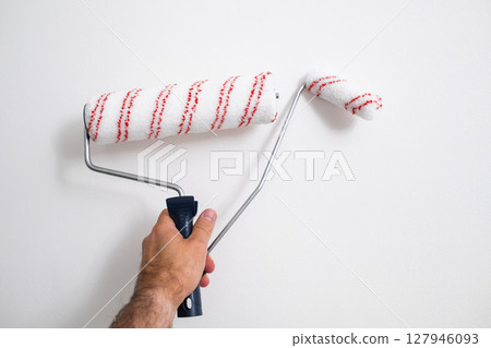 Close up of mans arm with medium sized and small paint rollers applying fresh coat of white paint. 127946093